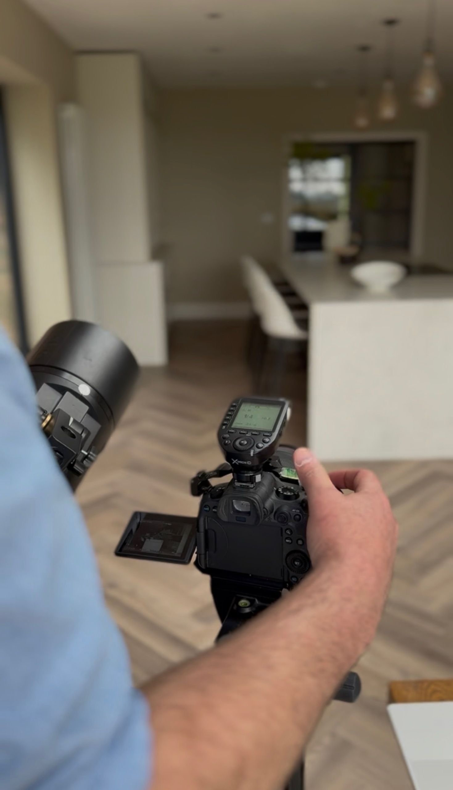 Behind-the-scenes view over a photographer&rsquo;s shoulder adjusting a professional camera mounted on a tripod. A modern kitchen with a white island is blurred in the background