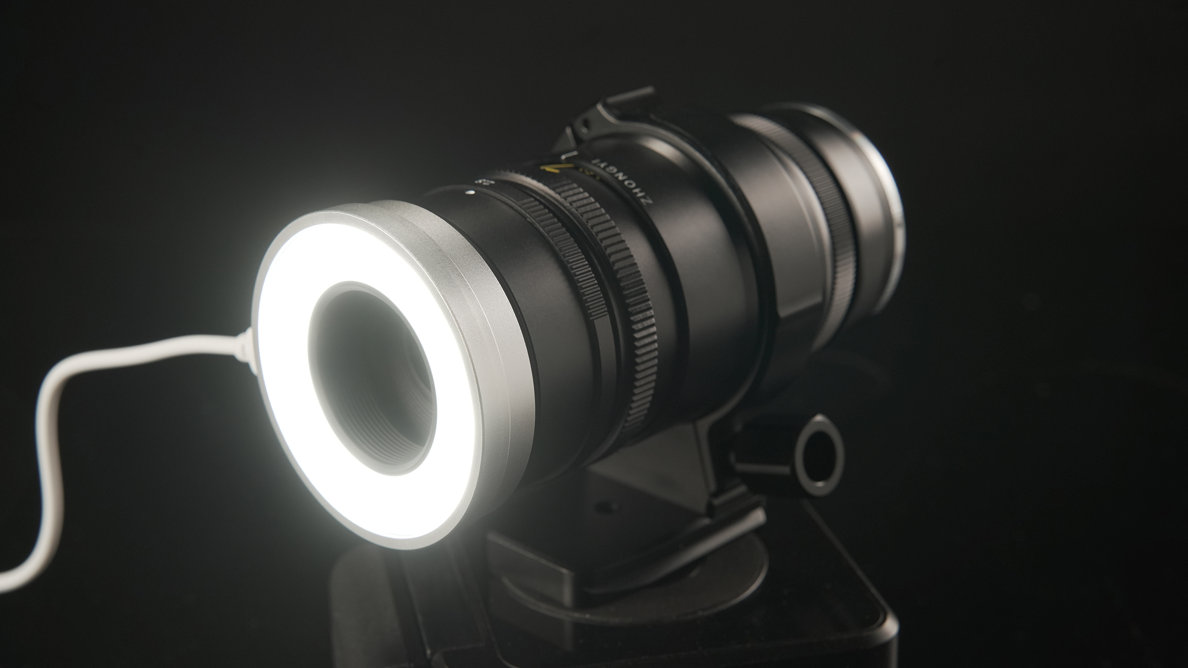 A close-up of a camera lens with a white LED ring light, resting on a black surface against a dark background