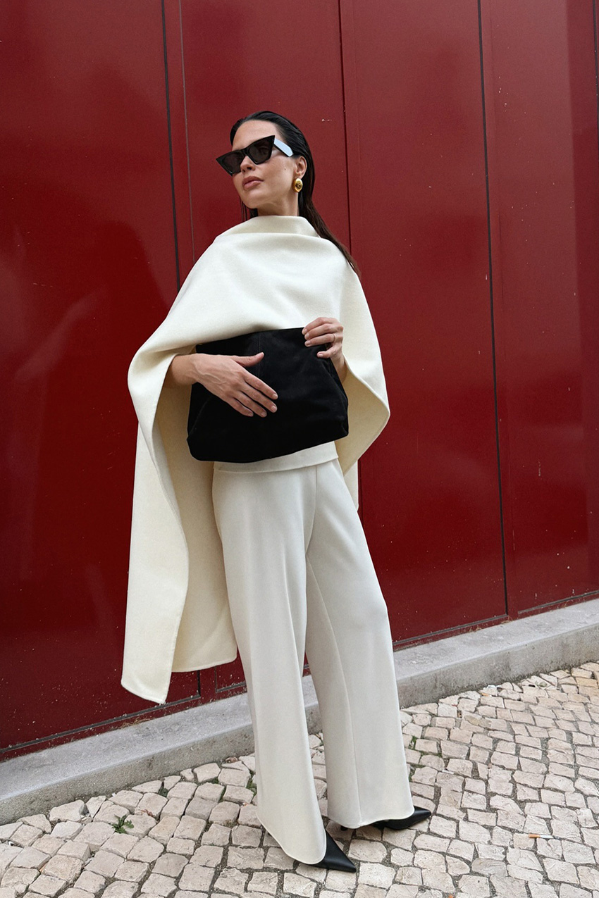 A woman standing on a sidewalk in front of a red wall wearing black sunglasses, gold earrings, a white cape over a white sweater, a black suede clutch, white trousers, and black pointed pumps.