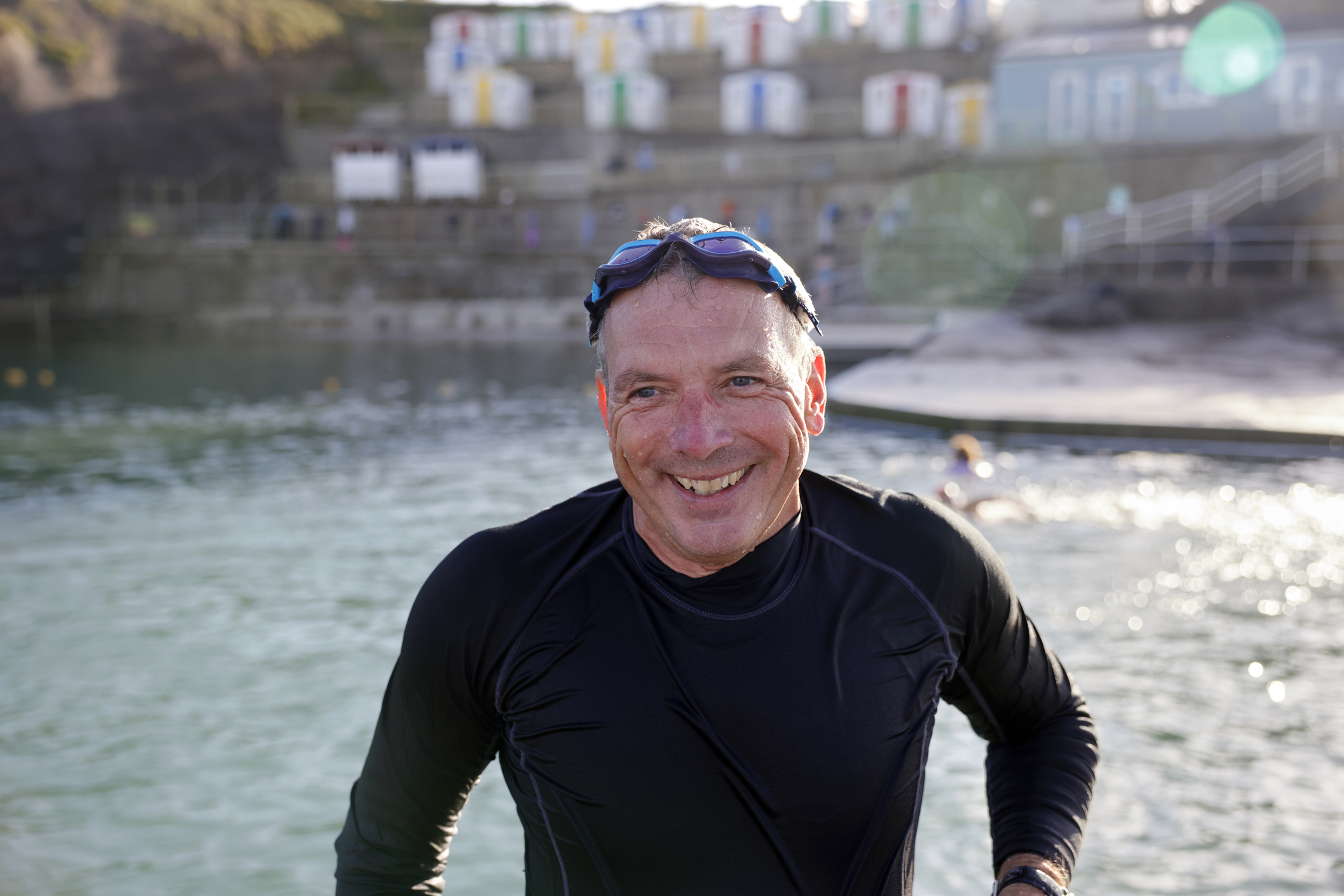 A man in his 50s emerges from a body of water wearing a wet suit. He is smiling and his goggles are perched on his head.