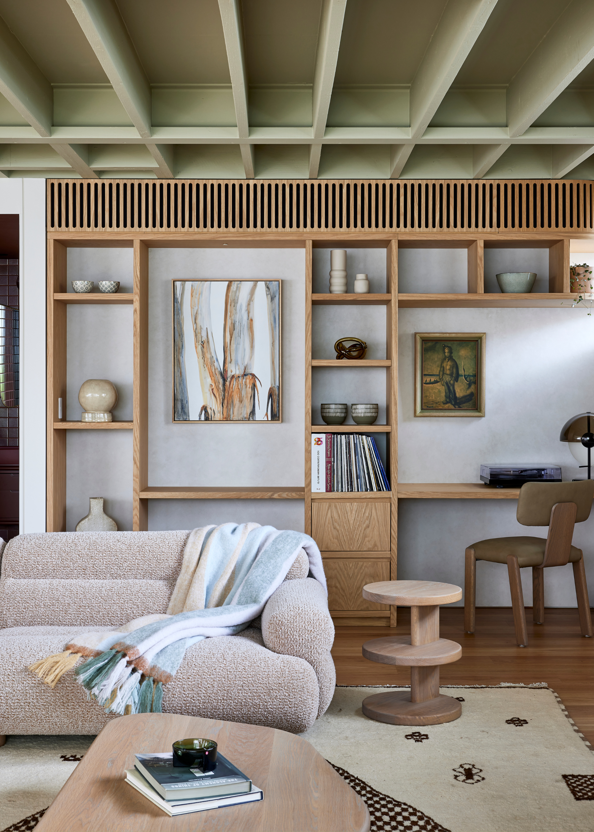A living room with a textured couch and a striped throw, a low wood coffee table with a candle and some books, a shelved wood stool, a large rug, a wood shelving unit, and a study space