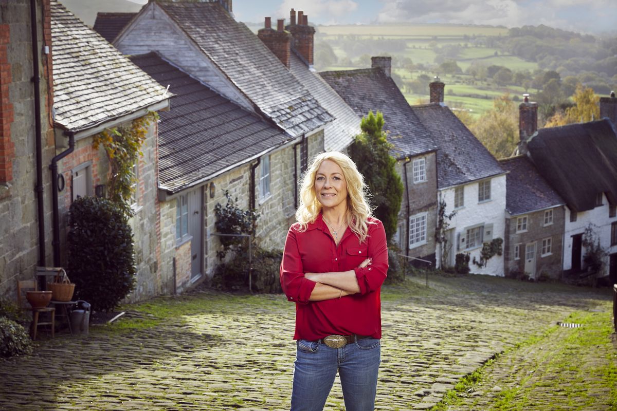 Sarah Beeny&#039;s New Country Lives: Sarah Beeny stands at the top of a cobbled country street which curves downhill and to our right behind her. The terraced street is full of quaint country cottages of different shapes, colours and sizes.