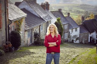 Sarah Beeny's New Country Lives: Sarah Beeny stands at the top of a cobbled country street which curves downhill and to our right behind her. The terraced street is full of quaint country cottages of different shapes, colours and sizes.
