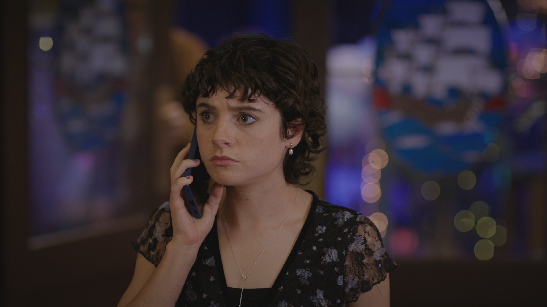 Indie at the wedding reception, wearing a dark, flowery dress and on the phone looking worried.