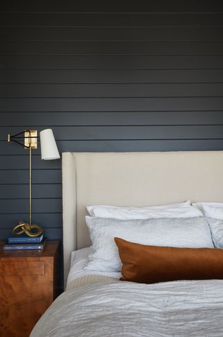 Image of a navy blue bedroom with a bed that has an off-white headboard and white bedding. There is a burnt orange bolster pillow on the bed and a warm wood side table. On the side table is a stack of blue books with a gold table lamp on top.