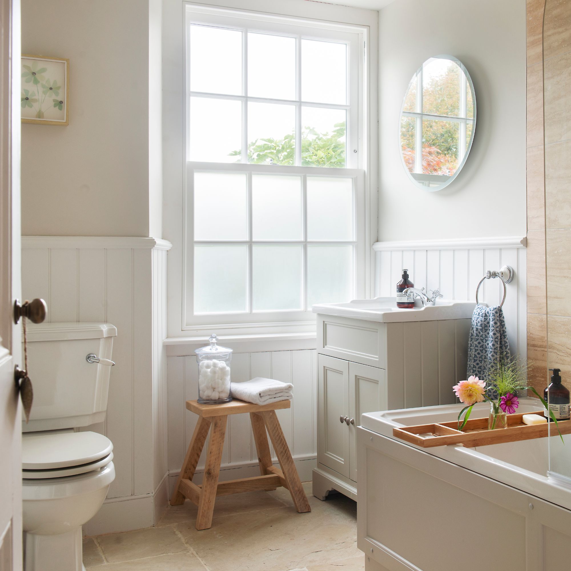 Bathroom in cottage with large window, white panelling, wooden stool and shaker-style tub and vanity