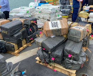 a haul of cocaine on pallets on a boat with authorities standing in between the bales
