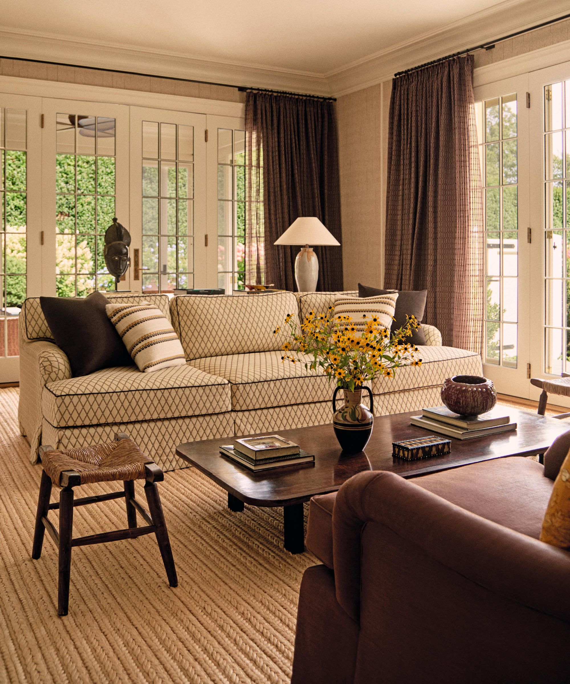 a cozy family room in the hamptons with textured wallpaper, french doors and textured beige rug layered with a lattice print sofa, antique wooden coffee table and plum chair