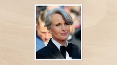 Andie MacDowell smiling with grey hair in a side-part updo and wearing a black tuxedo with oversized black bow tie, on a neutral background