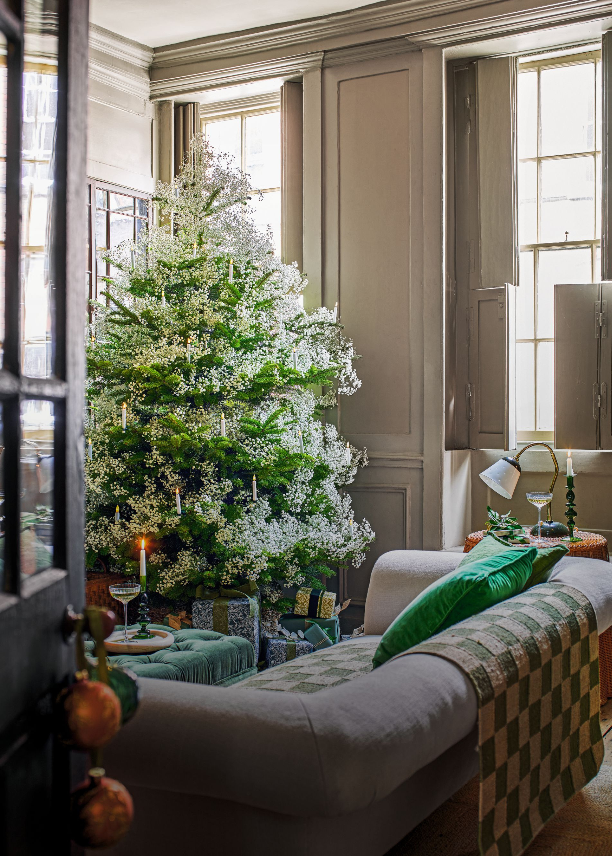 A large Christmas tree is adorned in white gypsophila with white candle decorations in a living room that has a checkered throw draped over the back of the sofa and baubles hang from the doorway handle