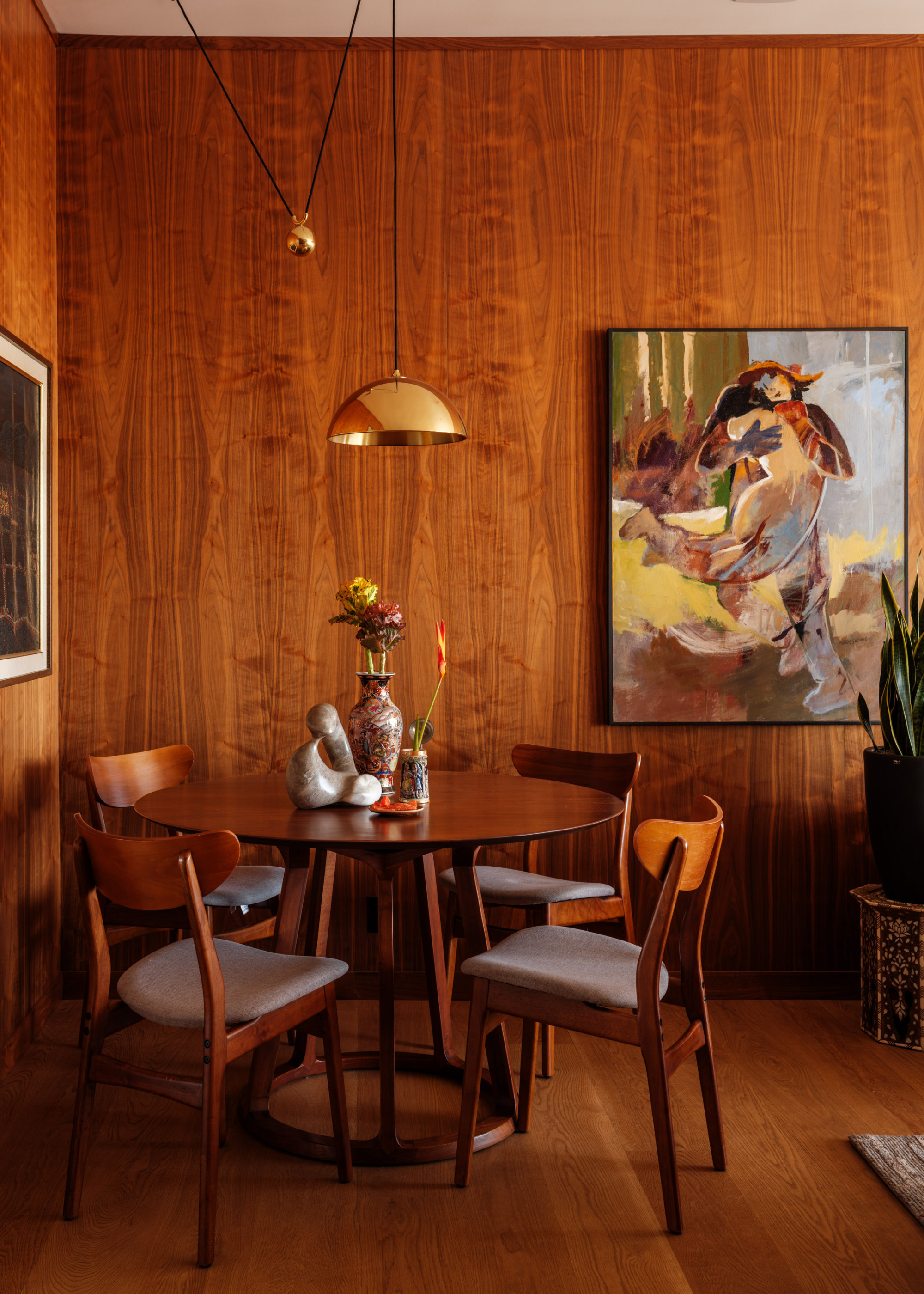 A wood dining room with a hanging brass pendant fixture, a round wood dining table with a set of chairs, a potted snake plant, and a hanging painting