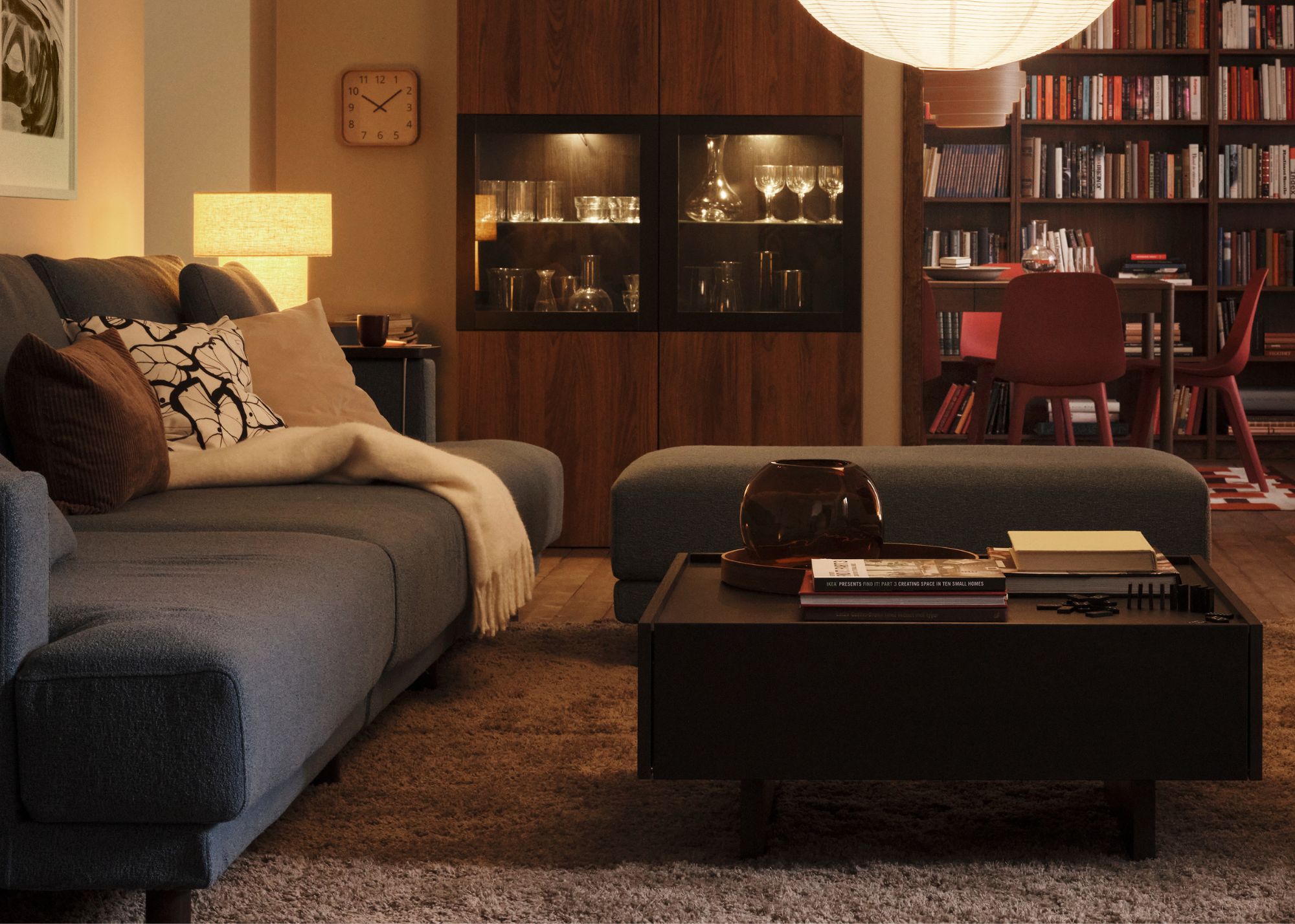 A cozy living room with a square dark wood coffee table, a gray sofa with cushions and a throw blanket and a gray pouf, with shelving and a dining table in the background and a lit table lamp