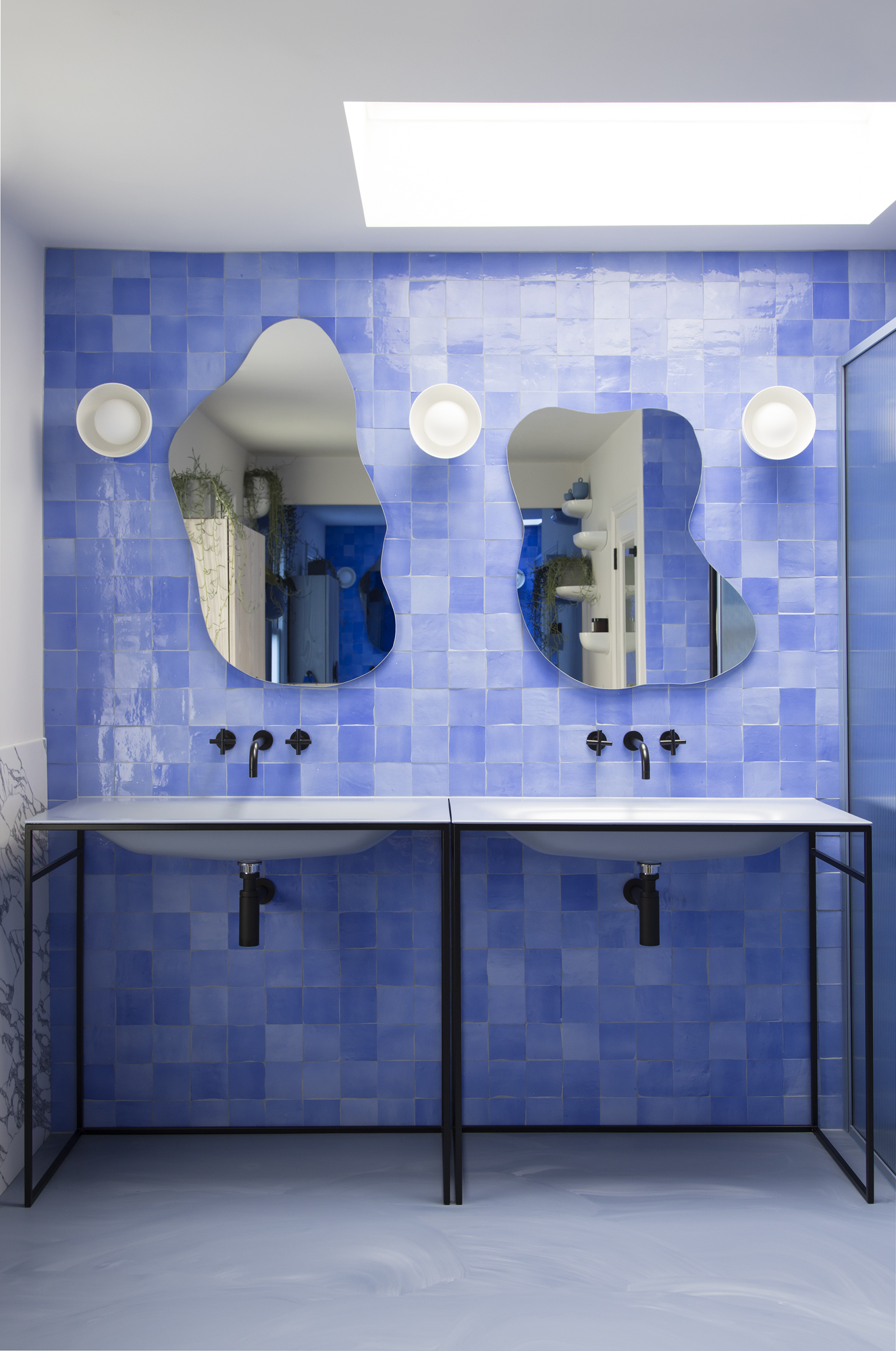 blue bathroom with bright blue zellige tiles, freestanding sinks, squiggly-shaped mirrors, wall sconces, and gray floors with skylight