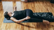 woman in black vest and leggings lying on her back on a black patterned yoga mat and wooden floor in a twist, facing away from the camera 