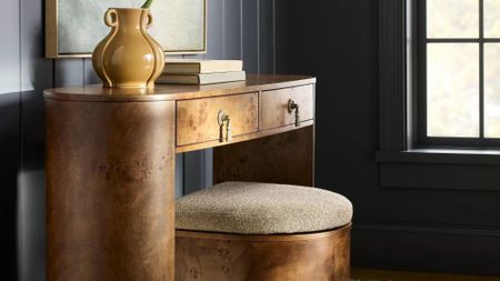 Shea McGee burled wood storage ottoman tucked under a coordinating burled wood desk inside of a navy wood paneled room with a large windowpane