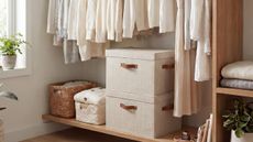 Two folding fabric storage boxes with lids at the bottom of a closet, with clothes hanging over the top