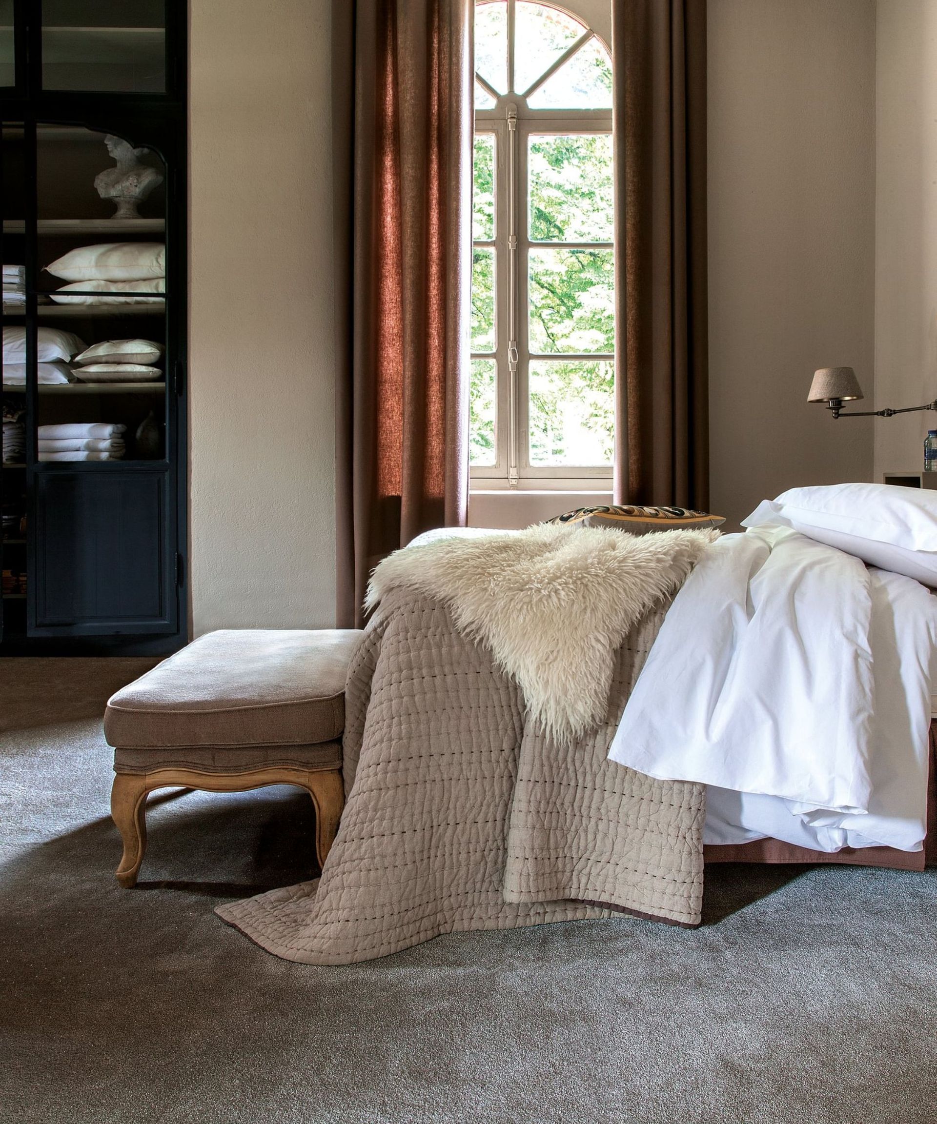 Bedroom with large arched window, grey carpet, blue shelves and bed with neutral bedding and stool