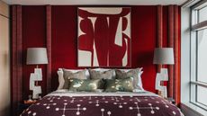 red bedroom with red wallpaper behind bed, red curtains, and red and white artwork hung over bed, sculptural white table lamps on either side of the bed dressed with white sheets, three bronze cushions, two decorative green cushions with swans, and a red and white blanket over the end of the bed