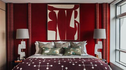 red bedroom with red wallpaper behind bed, red curtains, and red and white artwork hung over bed, sculptural white table lamps on either side of the bed dressed with white sheets, three bronze cushions, two decorative green cushions with swans, and a red and white blanket over the end of the bed