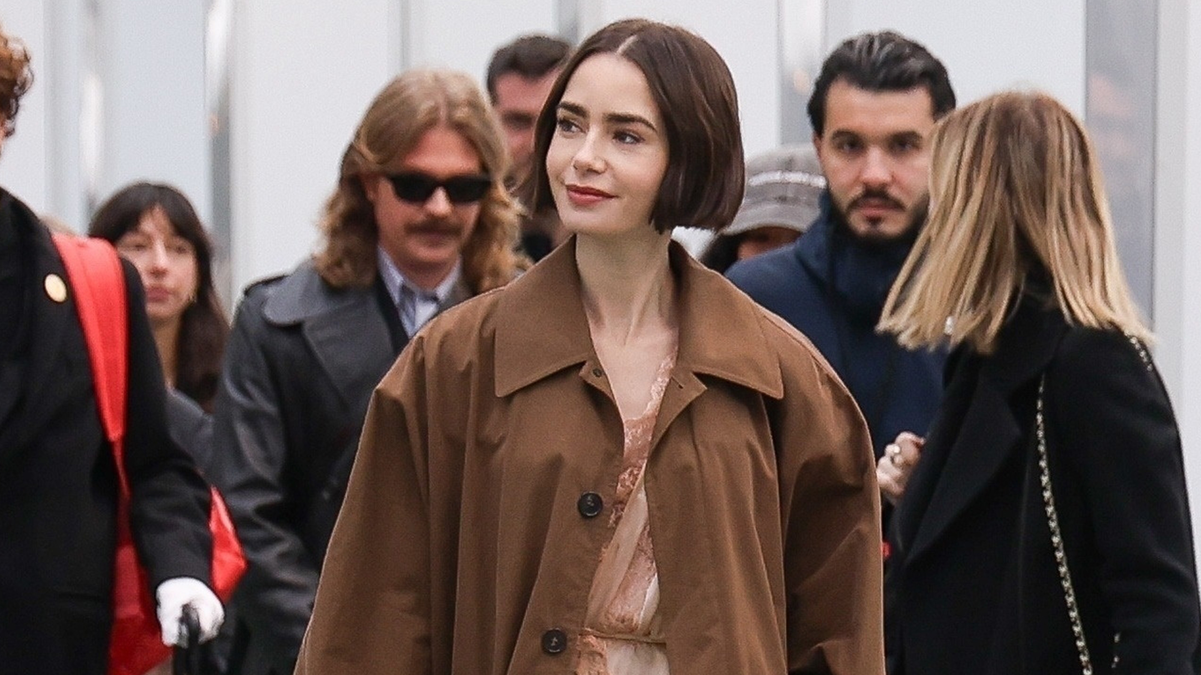 Lily Collins arrives at Gare d&rsquo;Austerlitz in Paris wearing a lingerie romper from YSL with a trench coat, tights, and heels, for Emily in Paris.