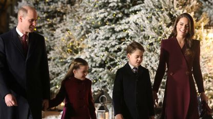 Prince William wears a black coat and Kate Middleton wears a dark red coat as they hold hands with their children Princess Charlotte and Prince George while walking in the snow