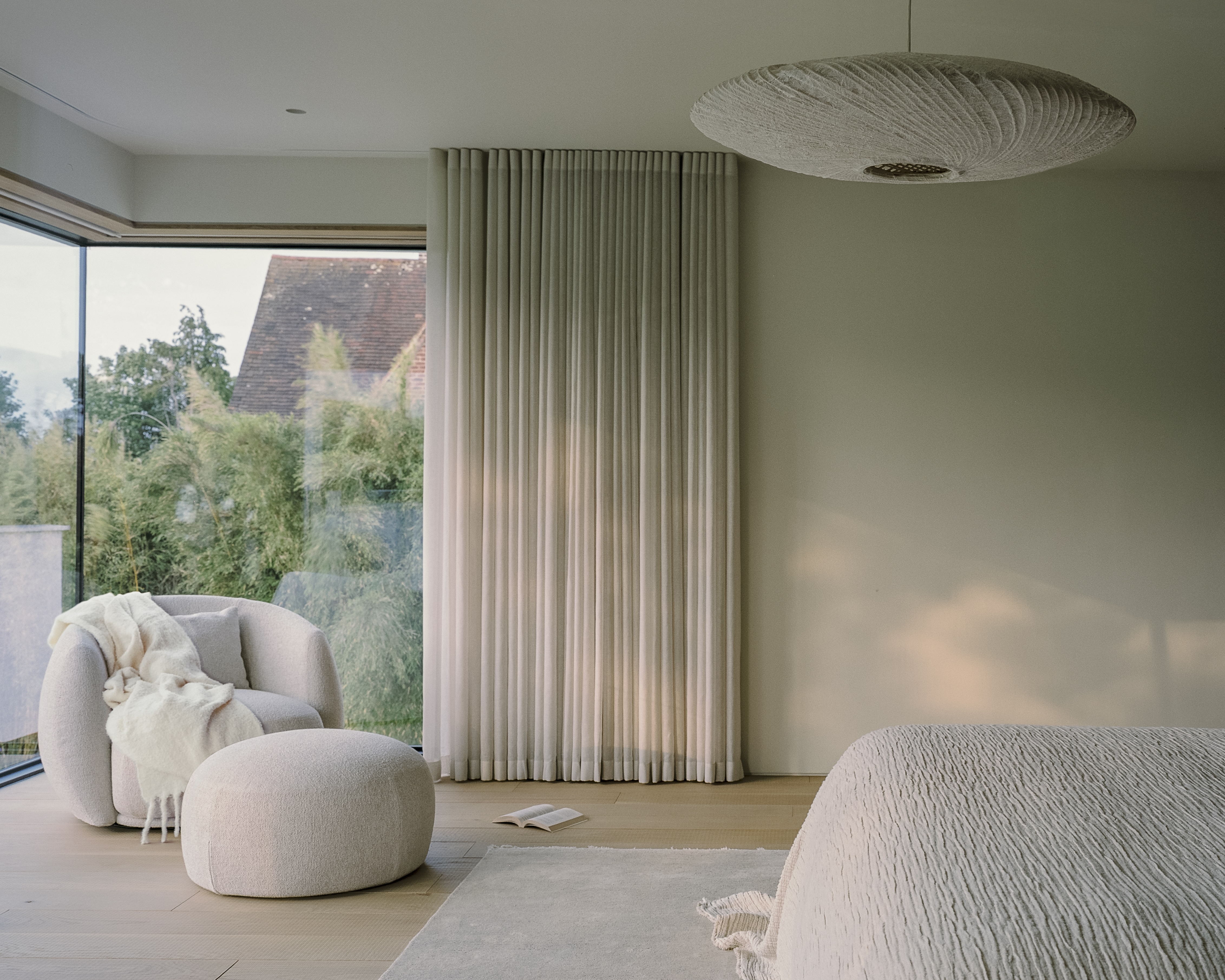 Contemporary bedroom with wave-fold curtains covering floor-to-ceiling windows, with a cream armchair and ottoman with a throw draped, timber floors, and cream bedcover