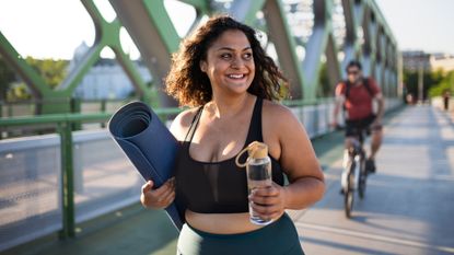 Best walking workout: Pictured here, a young woman walking outdoors on bridge in city carrying a yoga mat and a water bottle 