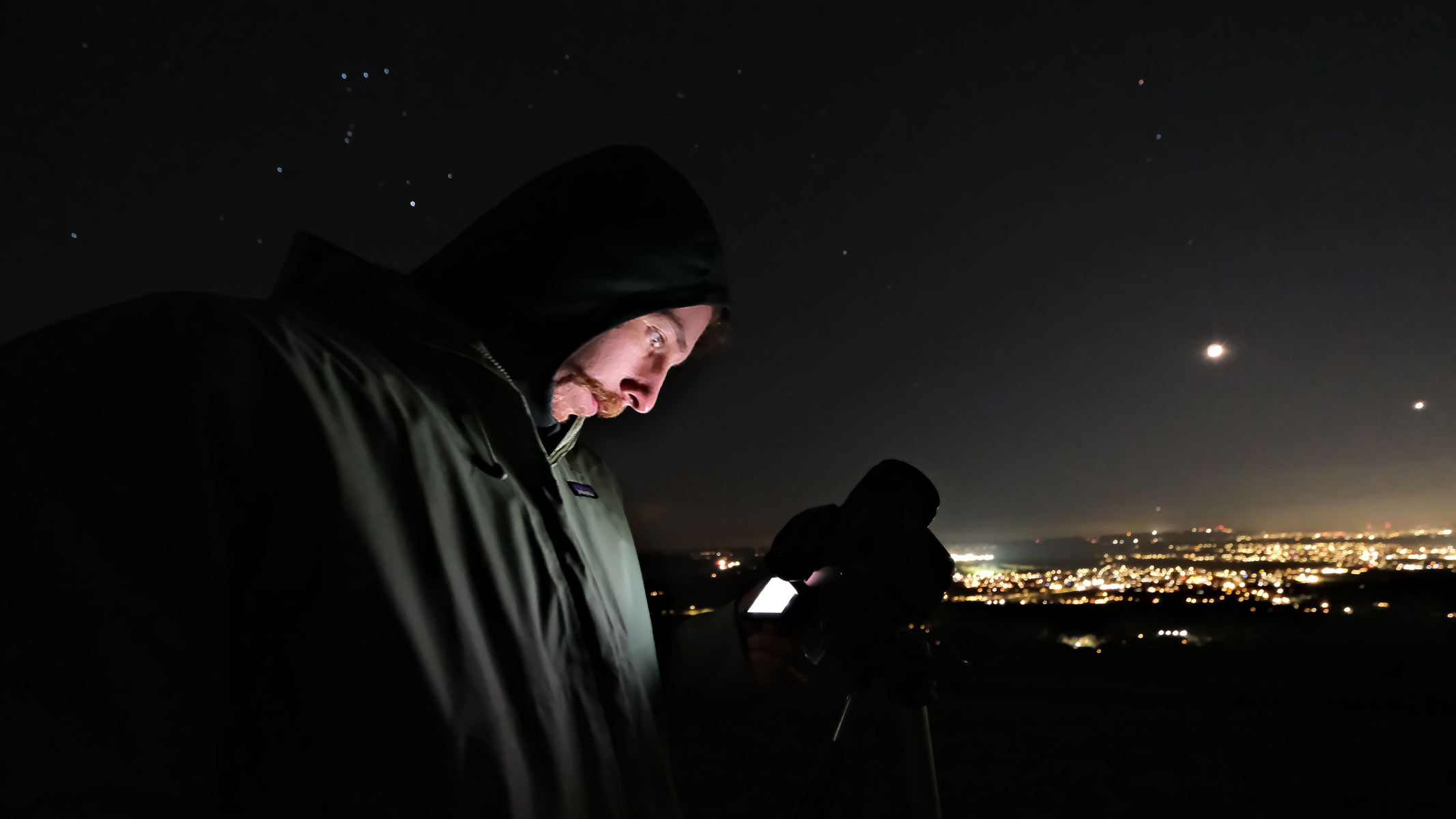 A man looking at a Fujifilm X-H2 with the stars, moon, Venus and city lights in the background