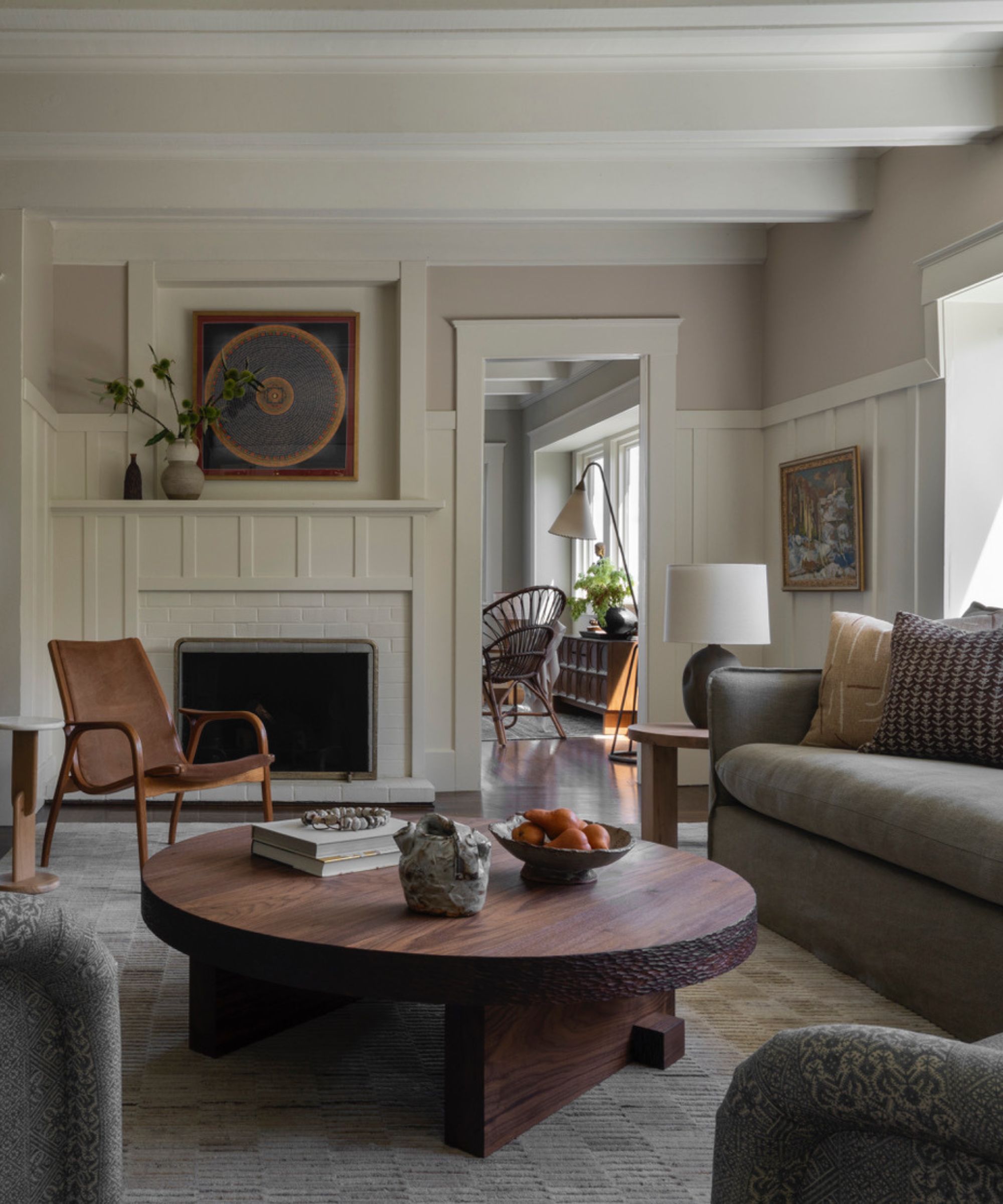 A cozy living room with lilac-gray walls, cream wall panelling and ceiling beams, a dark wood round coffee table with a textured rug, a gray sofa, and transitional decor.