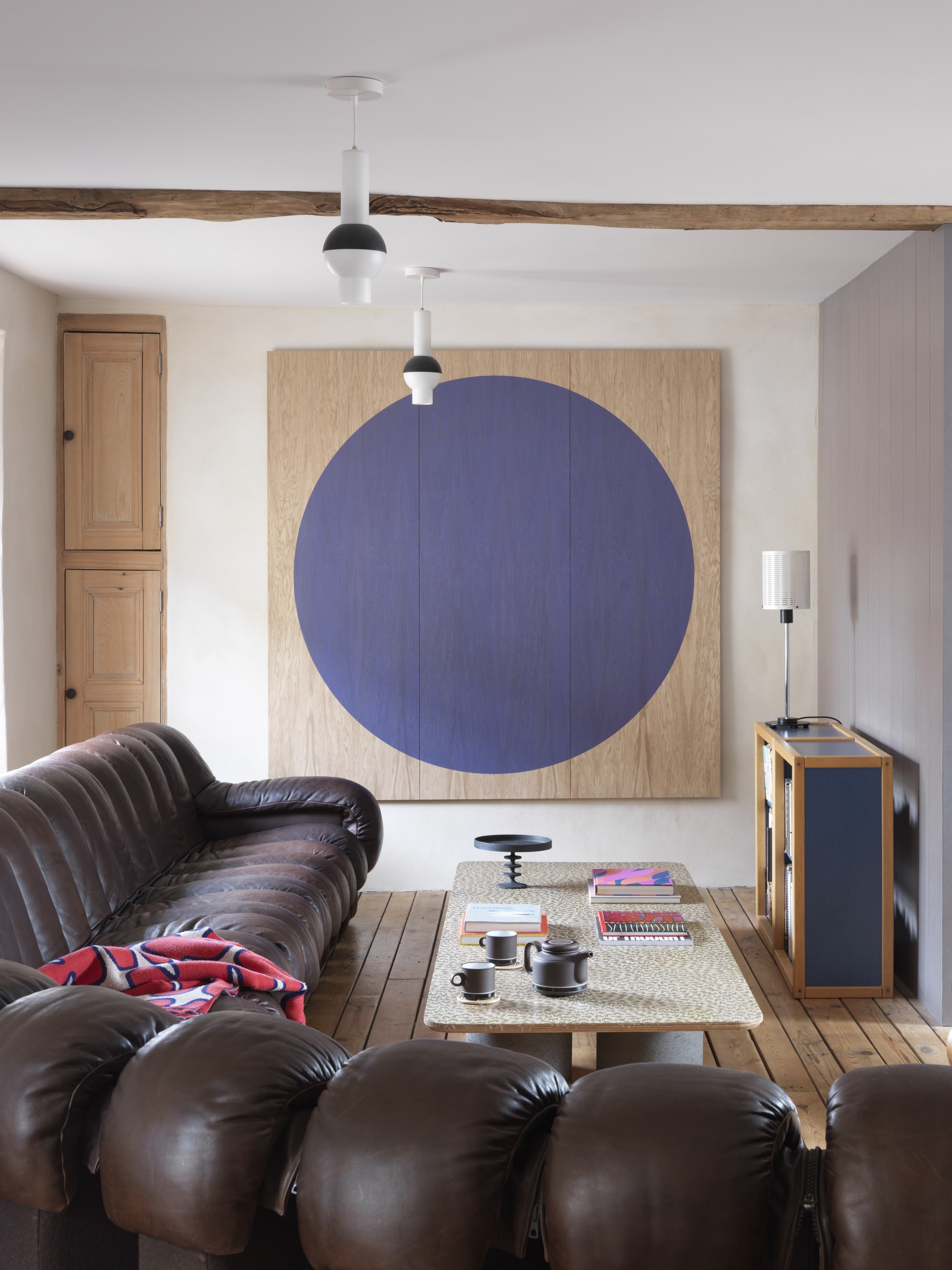 living room with timber ceiling beam, brown leather sofa, timber floorboards, timber artwork with painted blue circle, white walls, and coffee table styled with books and decor