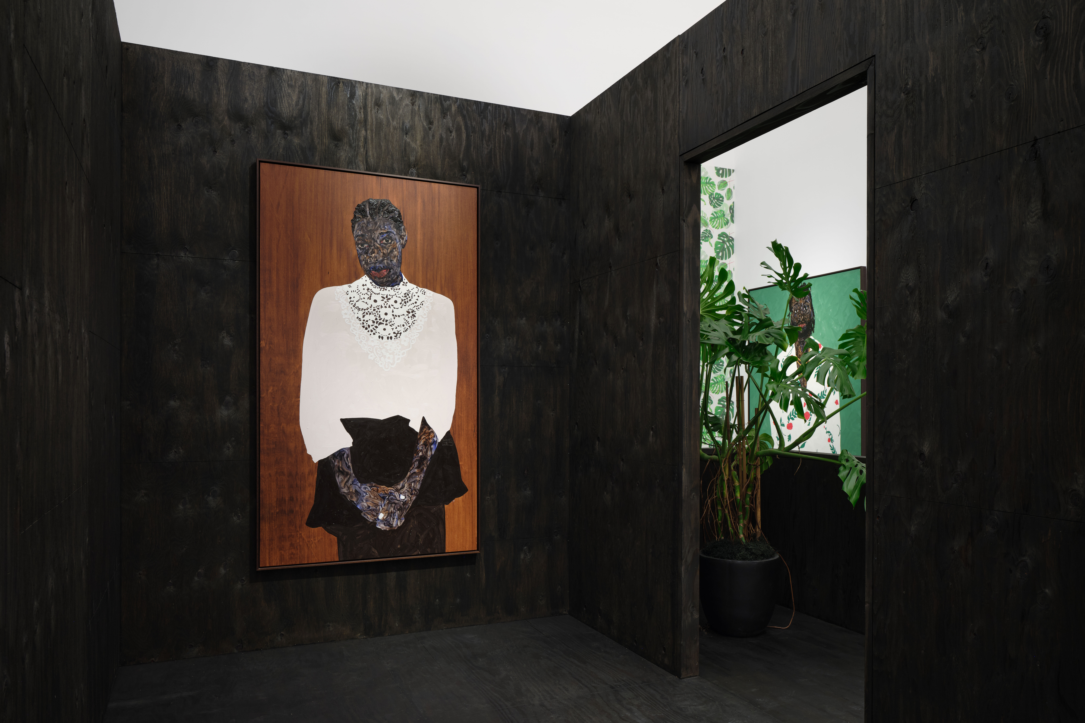 View of an art gallery installation with dark wood walls and multiple expressive portrait paintings. In the foreground, a large black metal structure with vertical bars frames additional paintings inside, while more portraits hang on the walls in the background.