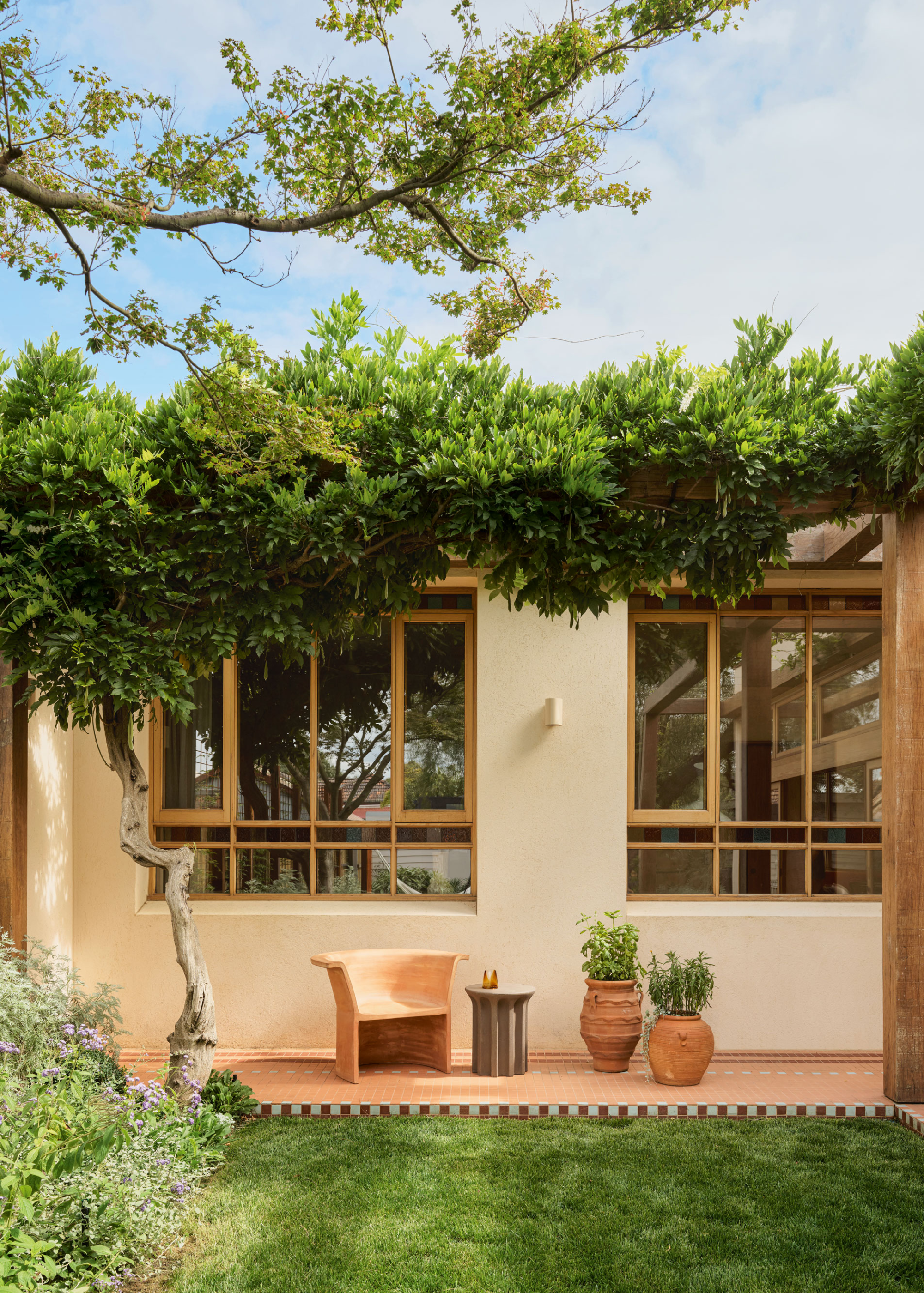 Neutral coloured exterior to the property and terracotta tile walkway with green lawn and plant pots in front. A pergola supports lush greenery at the full height of the windows and a tree sits to the left.
