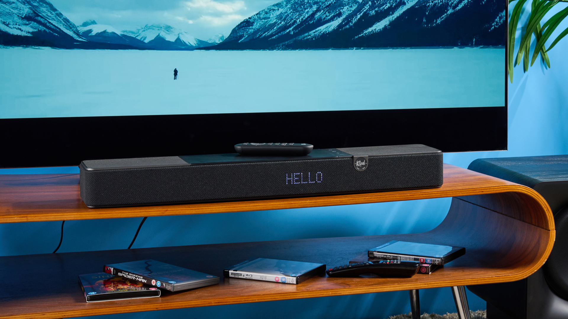 the klipsch flexus core 100 soundbar photographed on a wooden tv stand with a tv behind, showing the remote, buttons, and inputs
