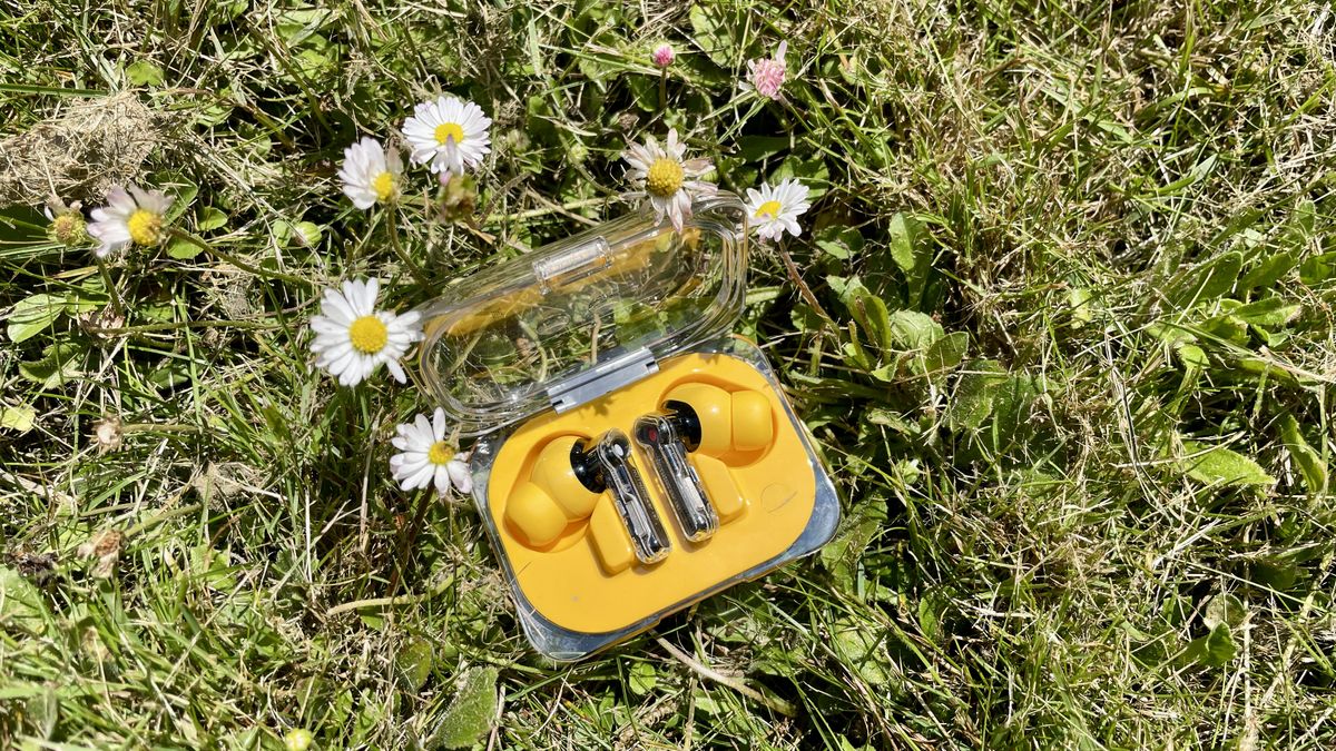 Nothing Ear (a) earbuds in yellow, on a grass lawn alongside yellow flowers, on a sunny day