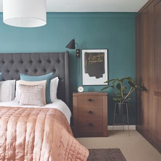Bedroom with bed, grey headboard and pink throw, next to bedside table and plant with navy blue wall behind