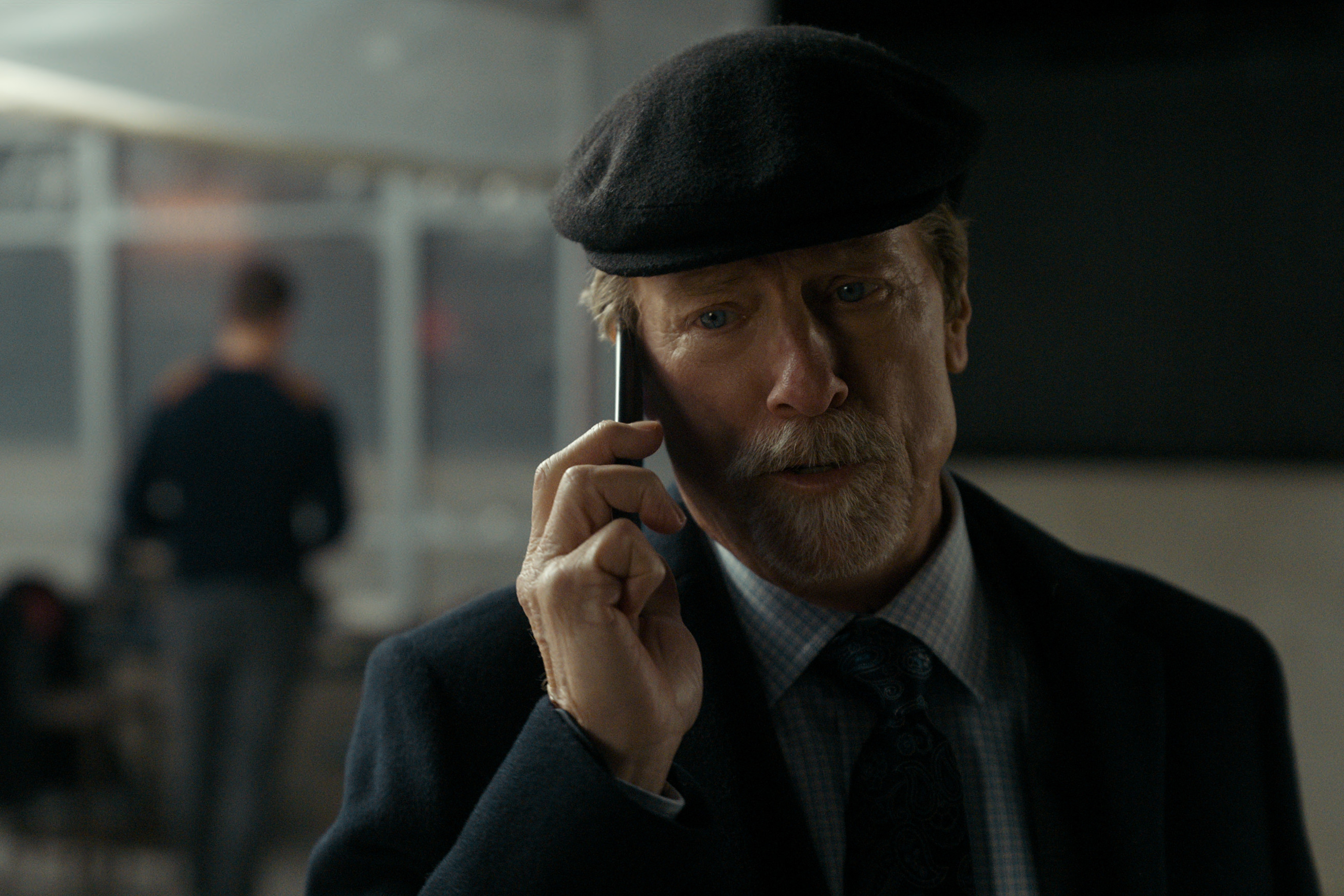 Louis Herthum as Jacob Monroe wearing a hat and talking on the phone on a balcony at night in a season 3 still of the night agent