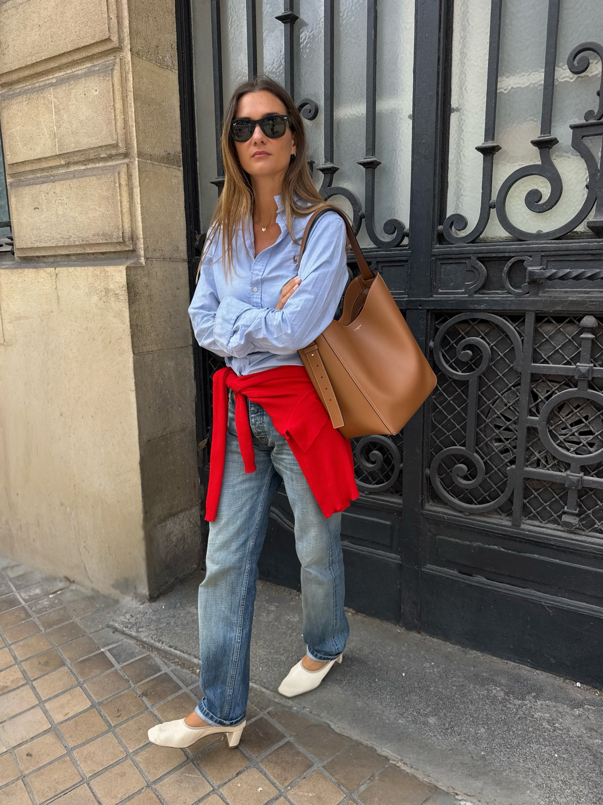 An image of an influencer in Paris wearing cream ballerina heels, an updated autumn shoe trend, with stovepipe jeans, a blue button-down and a red knit around her waist