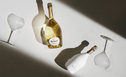 Bottles of Ruinart champagne, one with second skin sustainable packaging, on white table with glass