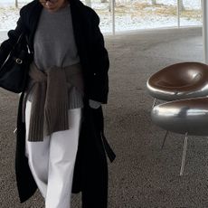 Woman wears a grey sweater with a sweater tied around her waist, white straight leg jeans and a black coat. 