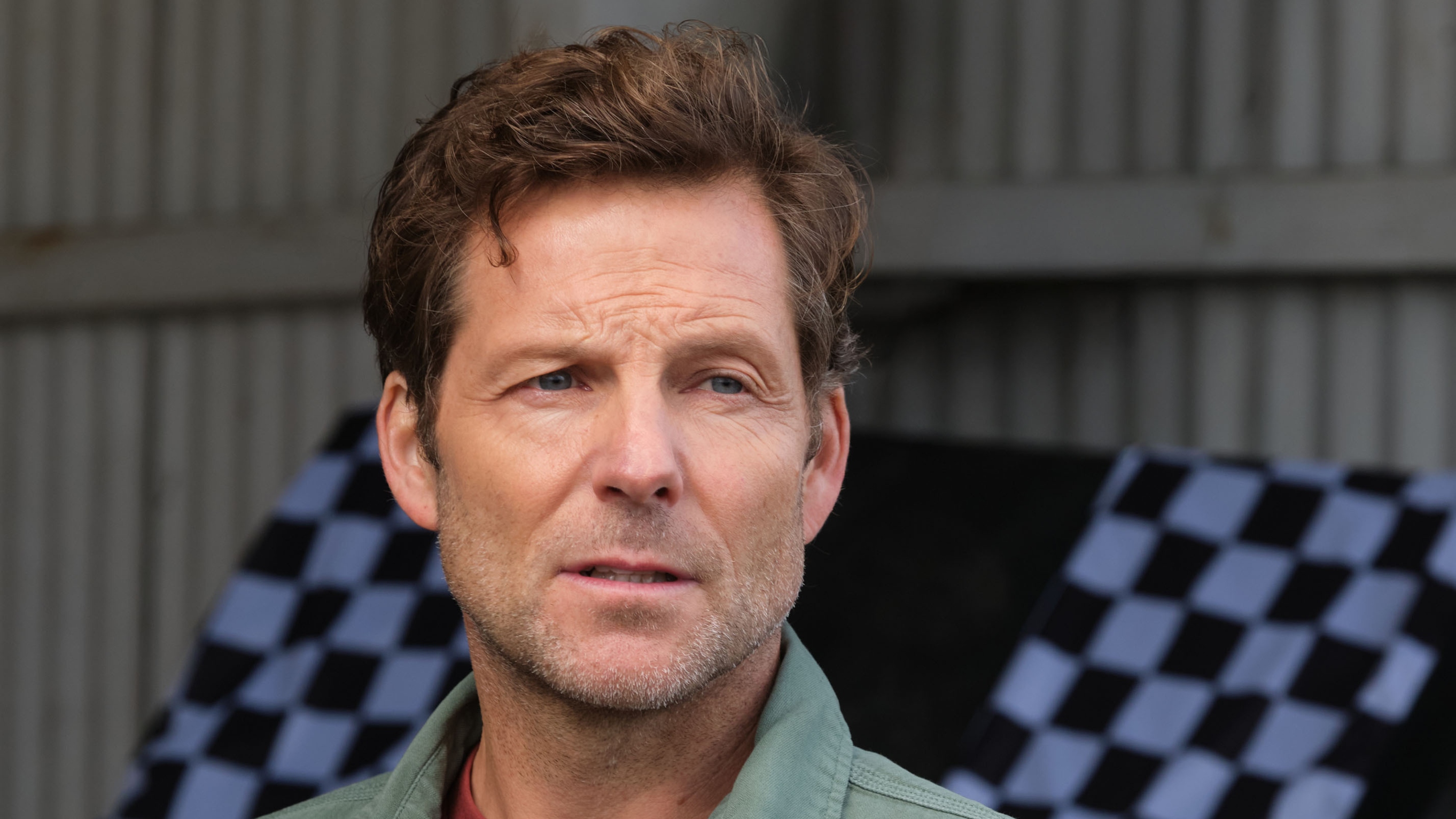 A close up of Archie Hughes (Jamie Bamber) inside an aircraft hanger, with some black-and-white chequered-flag style material behind him. He looks concerned.