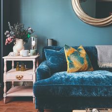 A blue-painted living room with a darker blue velvet sofa and a pink side table