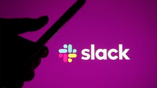 Slack logo and branding pictured on a purple screen with silhouetted hand holding smartphone in foreground.