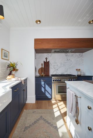 Image of a white kitchen with white marble countertops and backsplash. The cabinets are painted a navy blue color. The floors are hardwood and there is a tan runner rug in between the counter and the kitchen island.