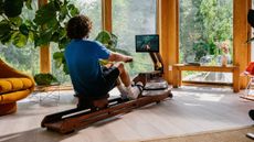 Man seen from behind rowing on wooden Ergatta rowing machine looking at screen in domestic setting