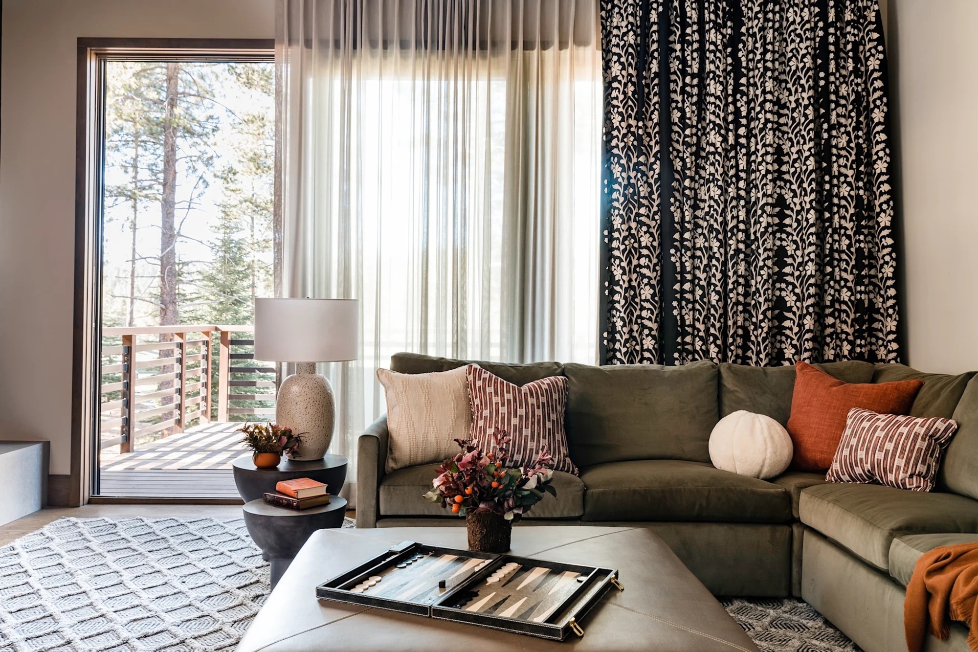 living room with patterned curtains hung over sheers, green l-shaped sofa with patterned cushions, leather ottoman coffee table with backgammon board and vase of flowers on it