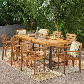 The Balfour 9pc Acacia Wood Dining Set on a outdoor rug, in front of greenery