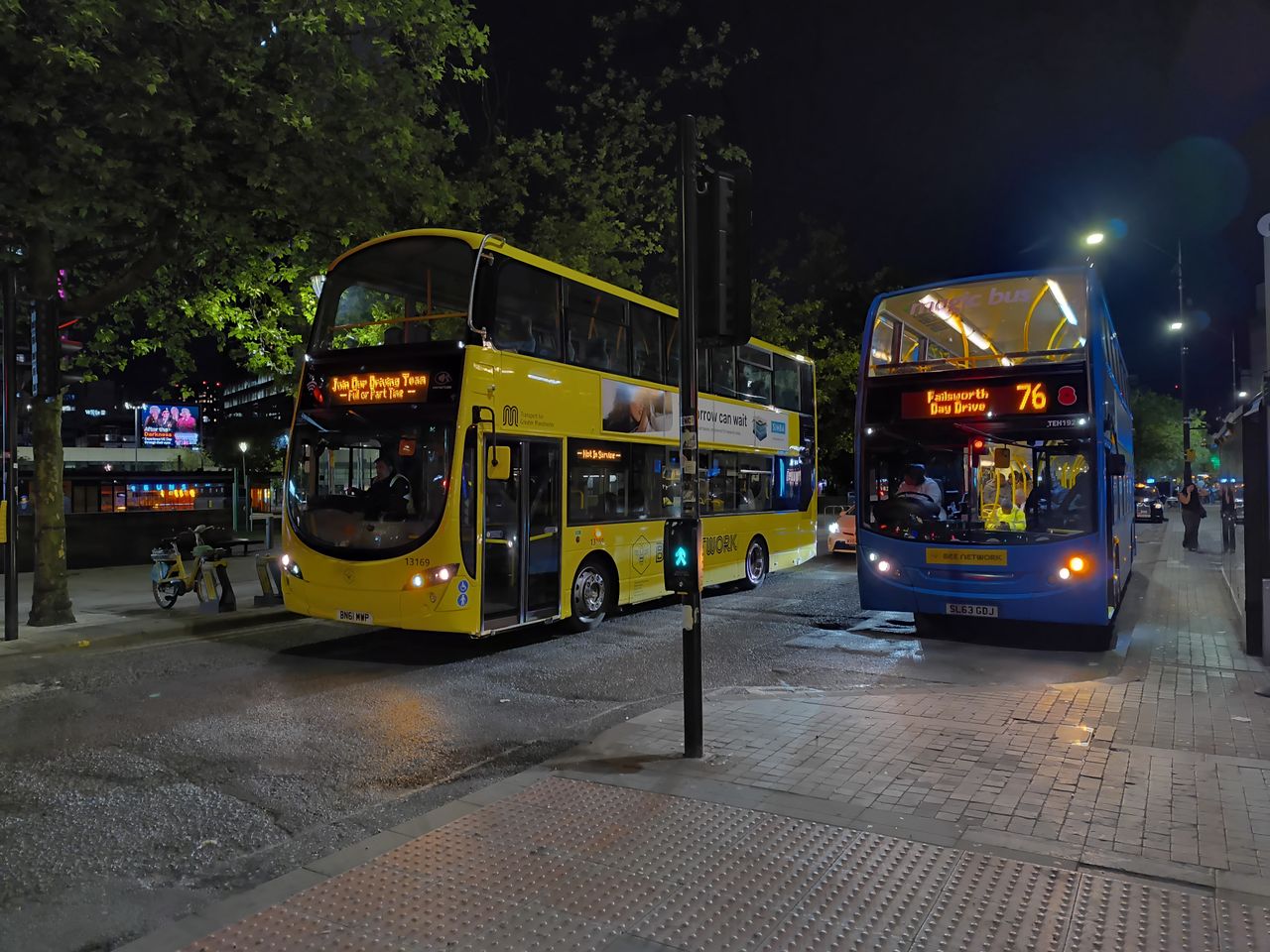 A nighttime street scene showing two double-decker buses stopped at what appears to be a bus stop.