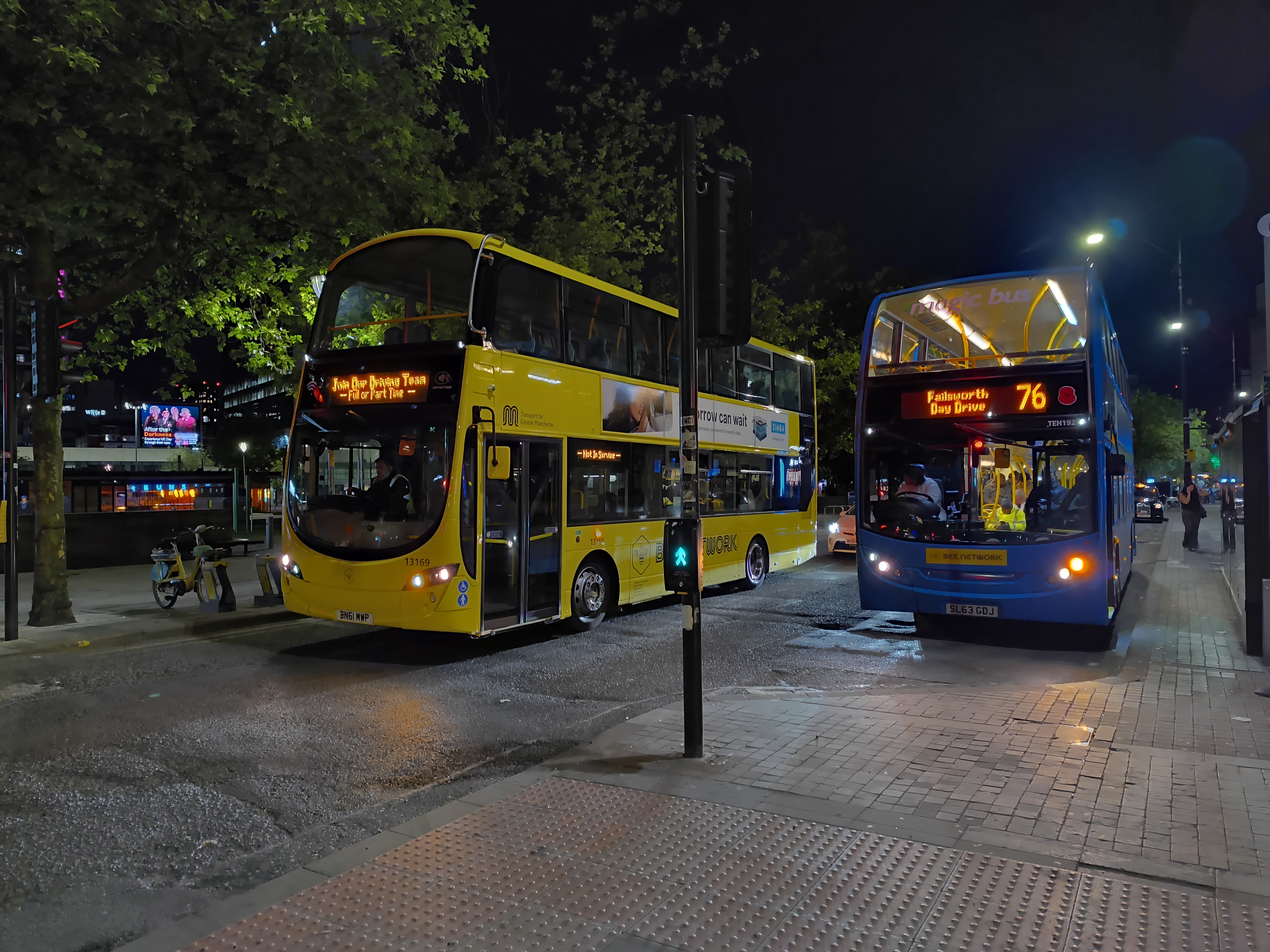 A nighttime street scene showing two double-decker buses stopped at what appears to be a bus stop.