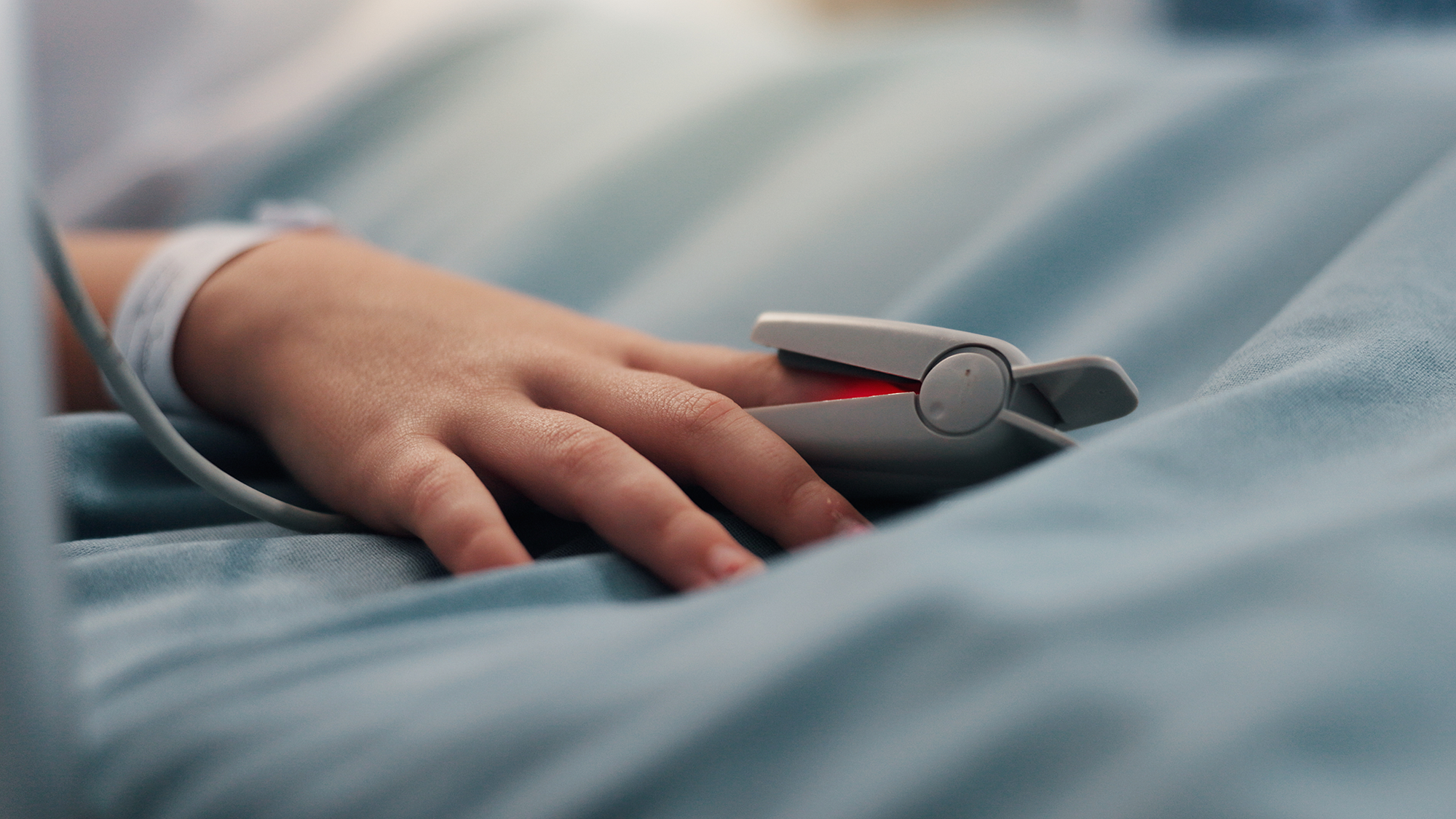 A close up of a child's hand resting on a hospital bed wearing a pulse-oximeter.