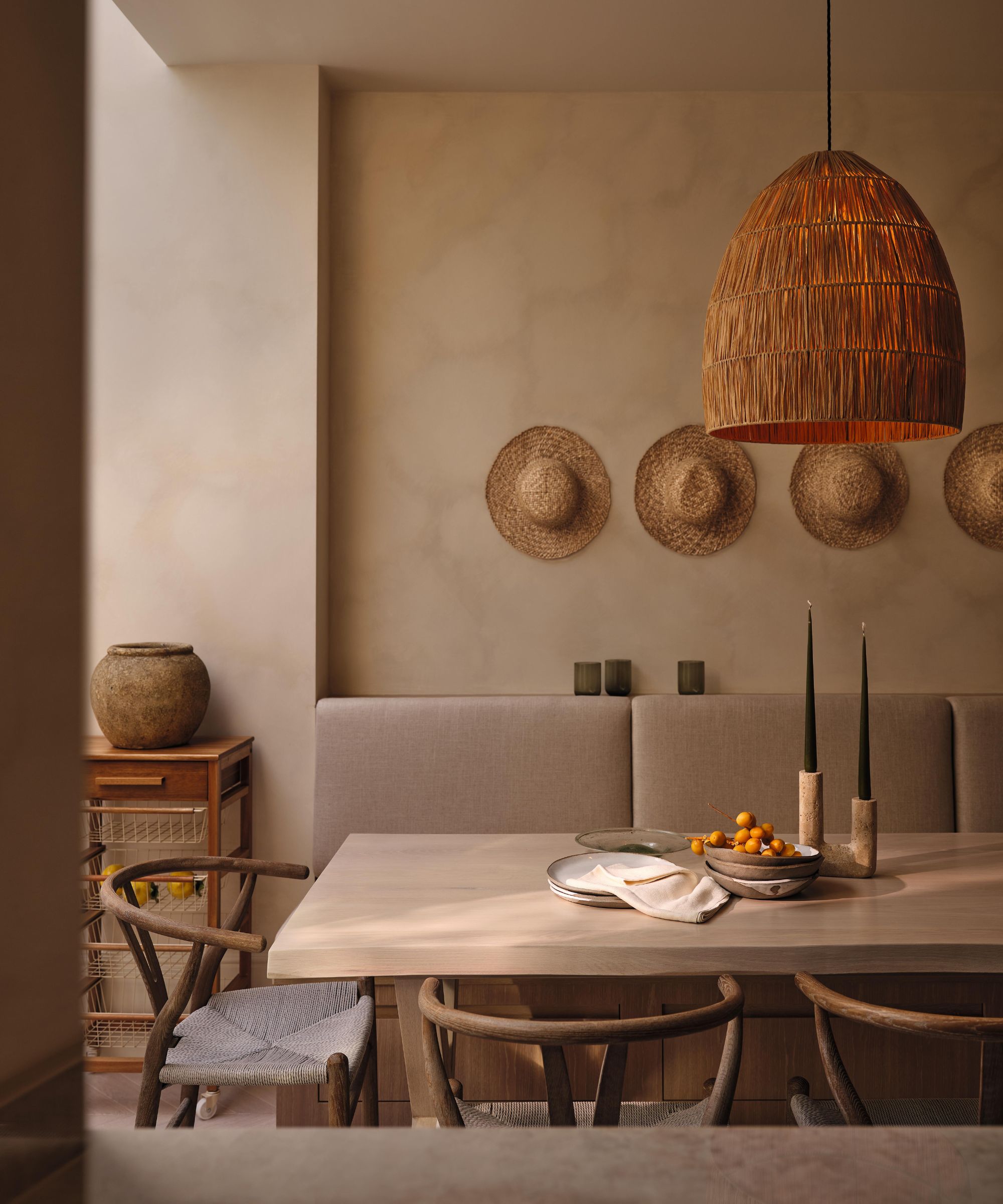 a limewashed kitchen dining area with a banquette seat, rattan hats on the wall, and a large woven pendant light
